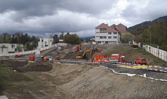 Eine Baustelle mit mehreren schweren Baufahrzeugen, einem Gebäude im Bau und einer Straße mit Fahrzeugen und Absperrungen.