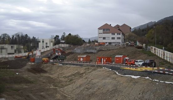 Baustelle mit Ausgrabung, Fahrzeugen und Maschinen. Ein großes Gebäude im Hintergrund. Bäume und Berge sind sichtbar.