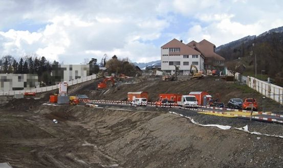 Eine Baustelle mit Fahrzeugen und Maschinen, einschließlich Baggern, auf einem Hang. Ein weißer Zaun begrenzt die Baustelle. Gebäude und Berge sind im Hintergrund.