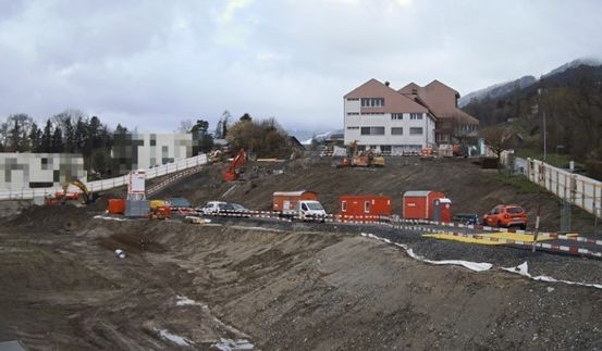 Eine Baustelle mit mehreren Fahrzeugen, Geräten und Gebäuden. Die Baustelle ist von einem Zaun umgeben, mit Bergen im Hintergrund.