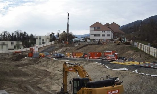 Eine Baustelle mit einem gelben Bagger, Absperrungen und einem Gebäude im Bau. Fahrzeuge und Maschinen sind anwesend, mit Bergen im Hintergrund.