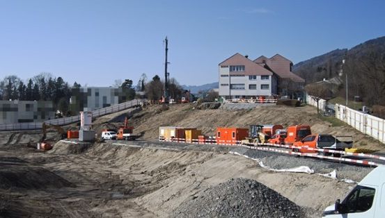 Eine Baustelle mit Baumaschinen, einem Lieferwagen, einem Haufen Erde und einem Haus in der Ferne unter einem blauen Himmel.