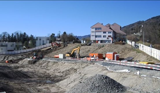 Eine Baustelle mit verschiedenen Baumaschinen, Erde und Kies. Gebäude und Bäume sind im Hintergrund sichtbar.