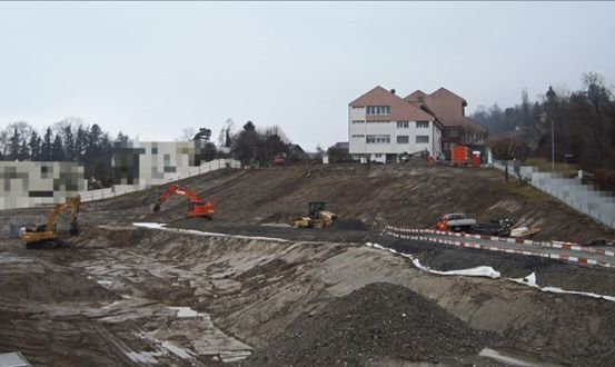 Ein Baustelle mit mehreren Baggern und Lastwagen ist zu sehen. Ein Gebäude im Hintergrund ist im Bau.