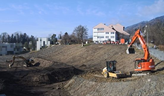 Ein Baugelände mit zwei orangefarbenen Baggern und einem gelben. Gebäude und Bäume im Hintergrund.