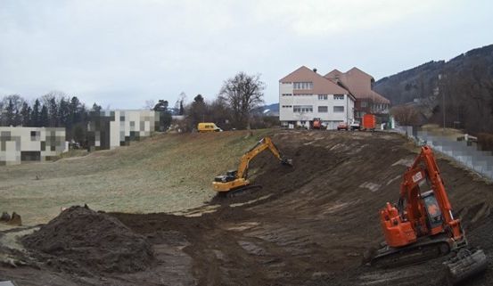 Ein Baugelände mit zwei Baggern auf einem Feld und einem großen Gebäude im Hintergrund. Der Himmel ist bewölkt.
