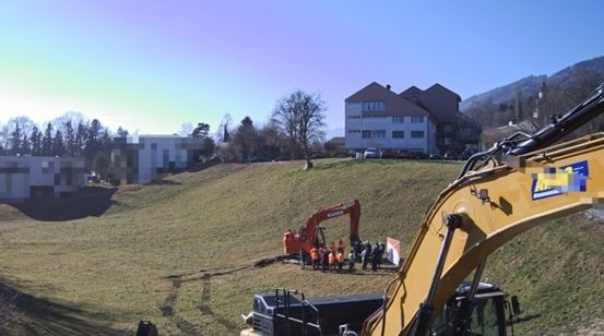 Eine Baustelle mit schwerem Gerät. Mehrere Arbeiter sind um einen roten Bagger versammelt. Im Hintergrund befinden sich Gebäude.