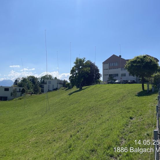 Ein Hügel mit grünem Gras und Bäumen, ein großes Gebäude und mehrere Antennen. Ein klarer blauer Himmel und eine Bergkette im Hintergrund. Das Datum und der Ort sind 14.05.25 und Balgach, respectively.