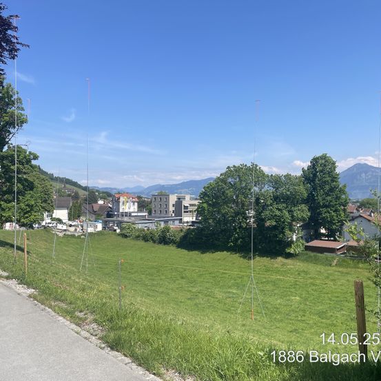 Ein Foto eines großen grünen Feldes mit hohen Bäumen und Bergen in der Ferne. Der Himmel ist blau mit einigen Wolken. Es ist 14:05:25 am 14. Mai 2025.