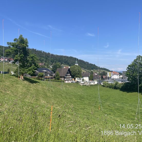 Ein grüner Grashügel mit mehreren hohen Antennen, Häusern und einem Berg im Hintergrund. Der Himmel ist klar und blau. Datum und Ort sind 14.05.25 1886 Balgach V.