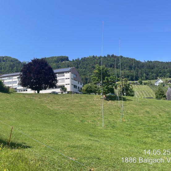 Ein offenes Grasfeld mit mehreren hohen Antennen und einem großen Gebäude mit Solarmodulen im Hintergrund. Datum und Ort sind unten angegeben.