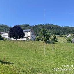 Ein offenes Grasfeld mit mehreren hohen Antennen und einem großen Gebäude mit Solarmodulen im Hintergrund. Datum und Ort sind unten angegeben.