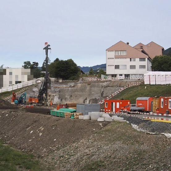 Ein Baugelände mit schweren Maschinen und Absperrungen. Im Hintergrund ist ein großes Gebäude sichtbar. Bäume und Berge sind in der Ferne.