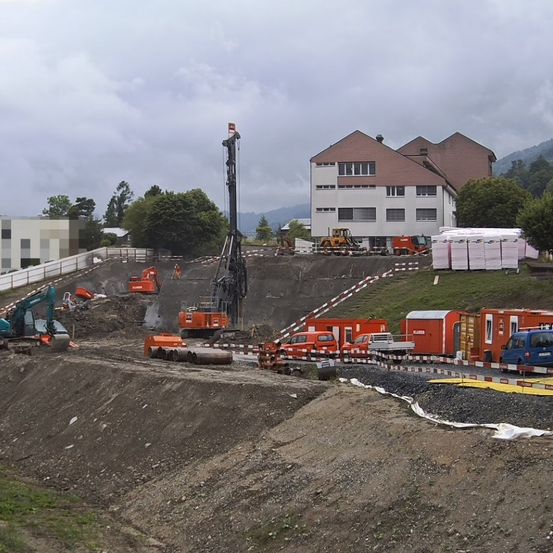 Eine Baustelle mit mehreren Baumaschinen, einschließlich einer hohen Bohrmaschine. Im Hintergrund befinden sich mehrere Gebäude und ein großer Erdhügel.