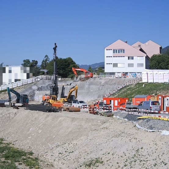 Eine Baustelle mit mehreren Baggern und Kränen an einem sonnigen Tag. Ein Gebäude und einige Bäume sind im Hintergrund sichtbar.