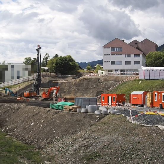 Eine Baustelle mit Maschinen, orangefarbenen mobilen Gebäuden und einem großen Haus im Hintergrund. Bäume und Berge sind in der Ferne sichtbar.