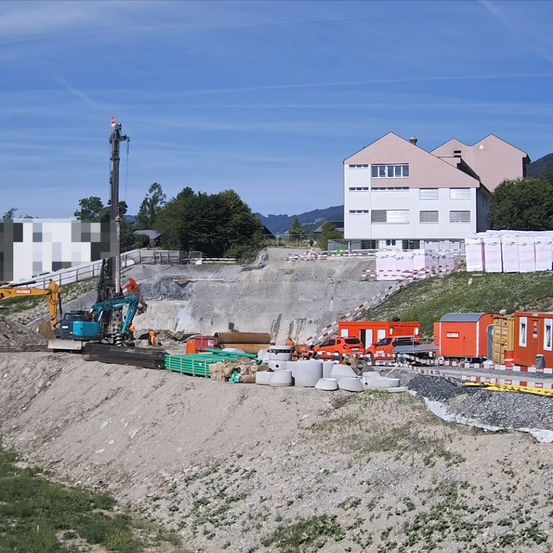 Ein Baugelände mit einem hohen Kran, schweren Maschinen und Arbeitern. Ein großes Gebäude im Hintergrund.