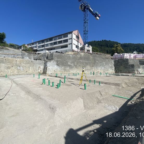 Ein Baustellengelände mit einem Kran, Messinstrumenten und grünen Pfosten an einem sonnigen Tag. Dahinter steht ein großes Gebäude. Datum und Uhrzeit: 18.06.2026, 10:00.