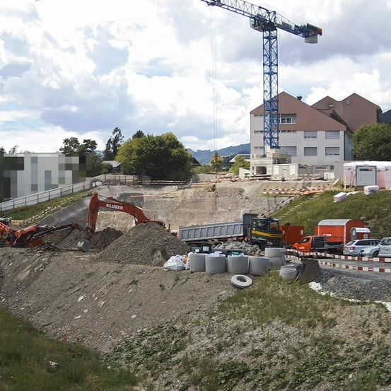 Eine Baustelle zeigt einen großen Bagger und einen Kran. Haufen von Erde und Baumaterialien sind verstreut. Hinter der Baustelle sind Gebäude und Bäume sichtbar.