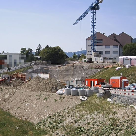 Eine Baustelle mit einem großen Kran, mehreren Gebäuden und verschiedenen Baumaterialien. Bäume und Berge sind im Hintergrund zu sehen.