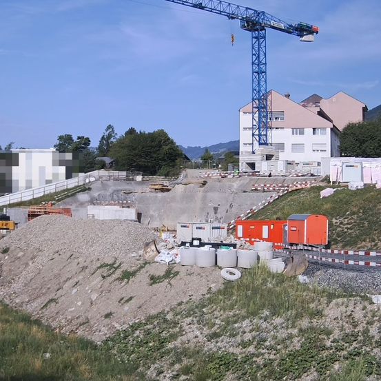 Eine Baustelle mit einem großen blauen Kran, umgeben von einem Zaun, mit einem im Bau befindlichen Gebäude im Hintergrund.