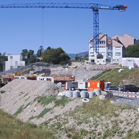Eine Baustelle mit einem hohen blauen Kran, mehreren geparkten Fahrzeugen und einem Haus im Hintergrund.