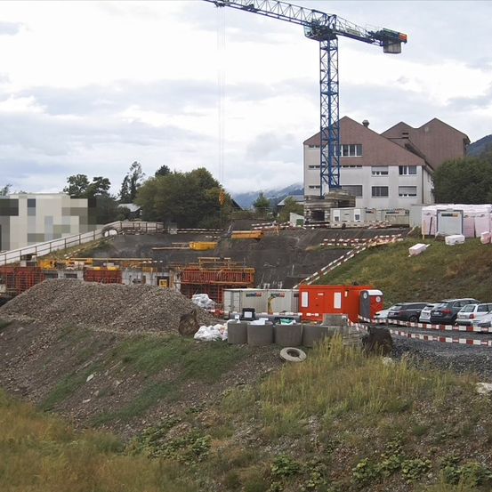 Eine Baustelle mit einem großen Kran, einem Gebäude im Bau und einem Haufen Kies. Bäume und Berge sind im Hintergrund.