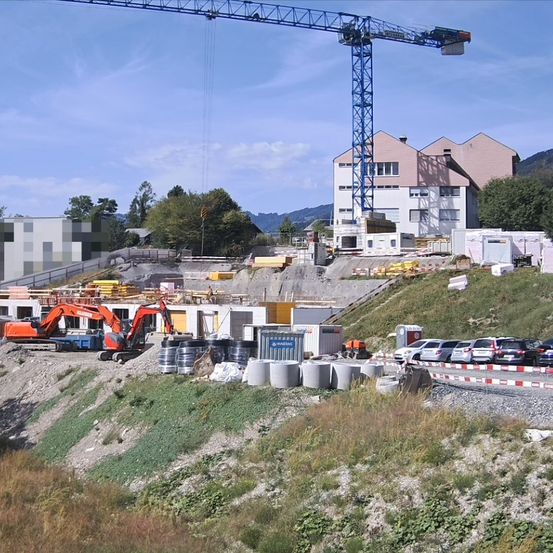 Eine Baustelle mit einem großen Kran, Maschinen und geparkten Autos. Gebäude und Bäume im Hintergrund.