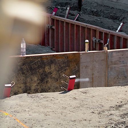 Ein Bauarbeiter in einem gelben Schutzhelm steht in der Nähe einer roten Betonwand mit Werkzeugen auf der Oberseite. Der Boden ist mit Erde bedeckt.