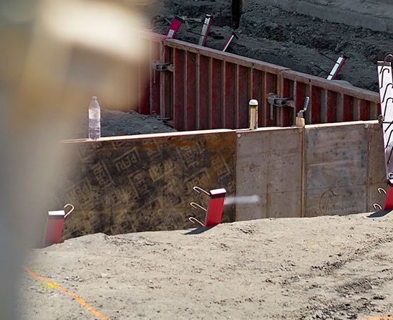 Ein Bauarbeiter in einem gelben Schutzhelm steht in der Nähe einer roten Betonwand mit Werkzeugen auf der Oberseite. Der Boden ist mit Erde bedeckt.
