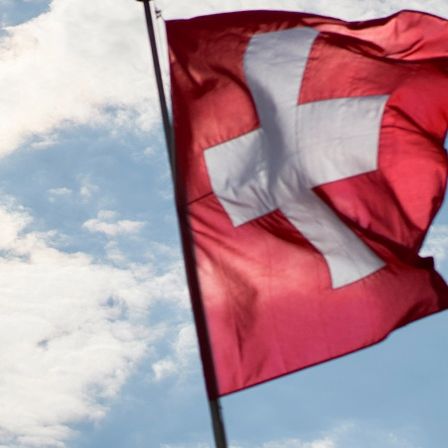 Eine rote Flagge mit einem weißen Kreuz in der Mitte weht im Wind gegen einen blauen Himmel mit weißen Wolken.