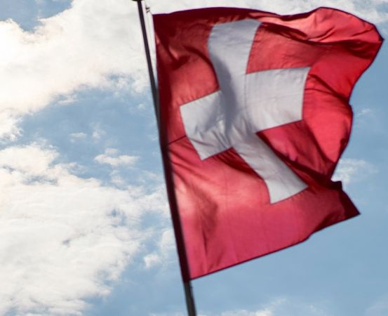 Eine rote Flagge mit einem weißen Kreuz in der Mitte weht im Wind gegen einen blauen Himmel mit weißen Wolken.