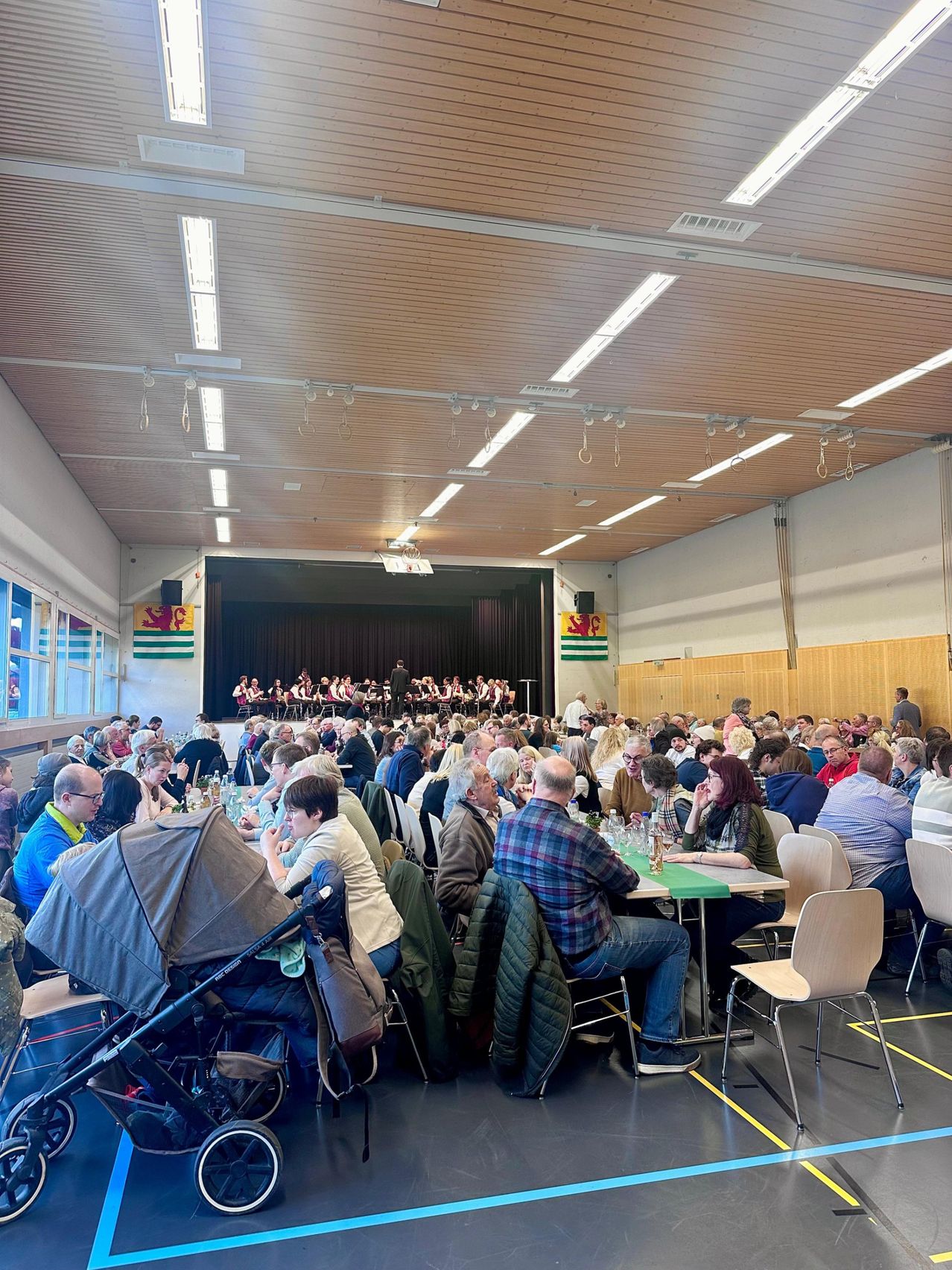 Eine große Veranstaltung im Innenbereich zeigt eine Menschenmenge an Tischen, eine Bühne mit einer Band, die spielt, und Flaggen an den Wänden. Die Decke ist mit Lichtern geschmückt.