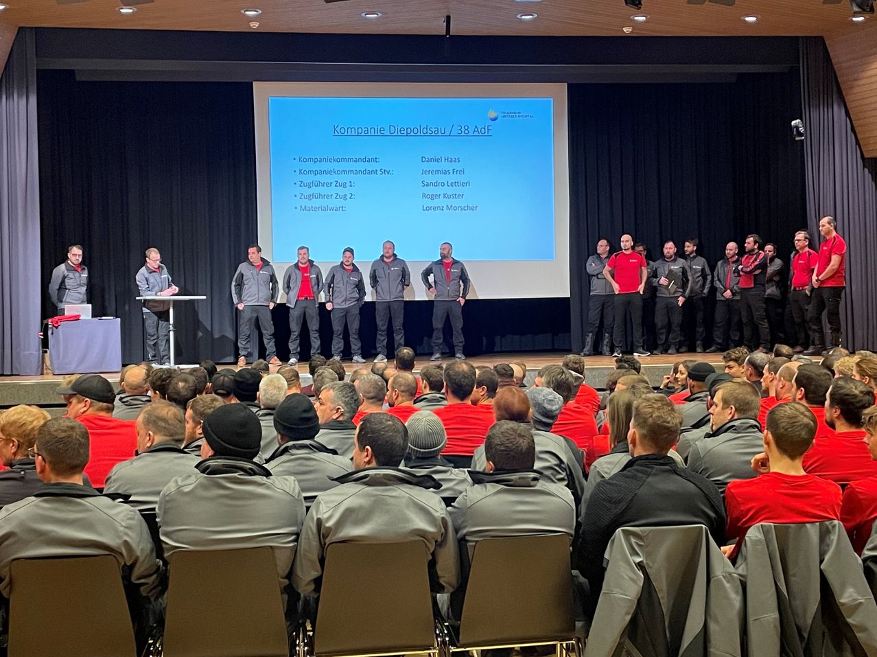 Eine Gruppe von Männern steht auf einer Bühne mit einer großen Leinwand dahinter, die 'Kompanie Diepoldsau / 38 Adf' zeigt. Sie tragen Uniformen und Mützen und nehmen wahrscheinlich an einer Präsentation teil. Ein Publikum davor beobachtet aufmerksam.