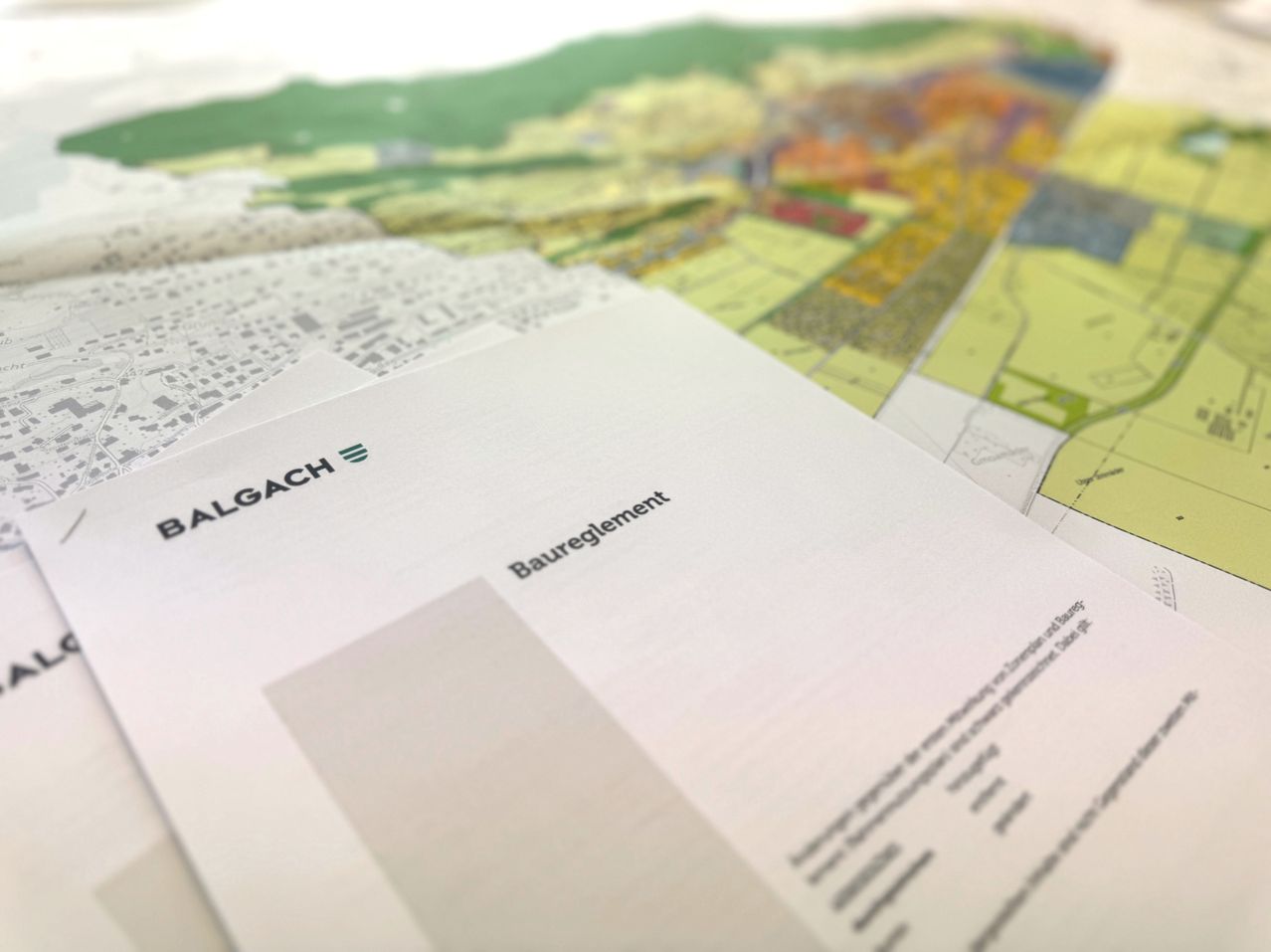 Ein weißes Dokument mit dem Wort 'Balgach' liegt auf einer Karte mit verschiedenen Farben und Markierungen. Die Karte zeigt verschiedene Bereiche, darunter grüne, gelbe und orange Zonen. Das Wort 'Bauregelung' ist auf dem Dokument sichtbar.