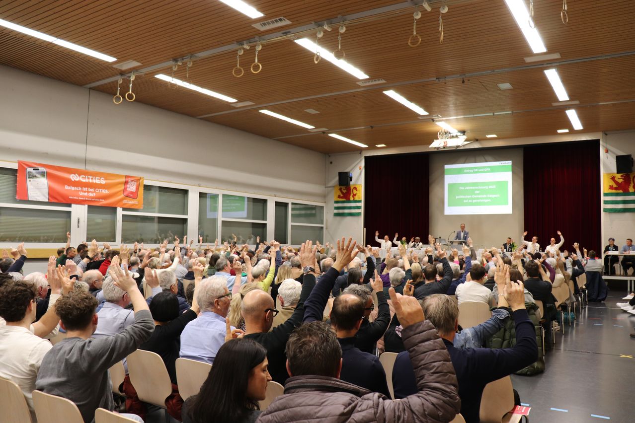 Audience raising hands in a hall, likely during a meeting. Flags and a projector screen with a presentation are visible.