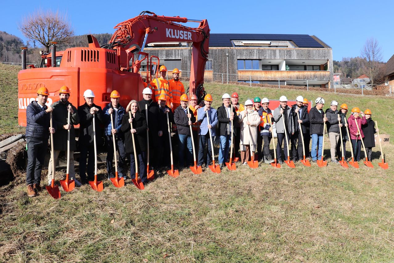 A group of people in helmets and coats stand in a line holding orange shovels in front of a red Kluser excavator and a building with solar panels.