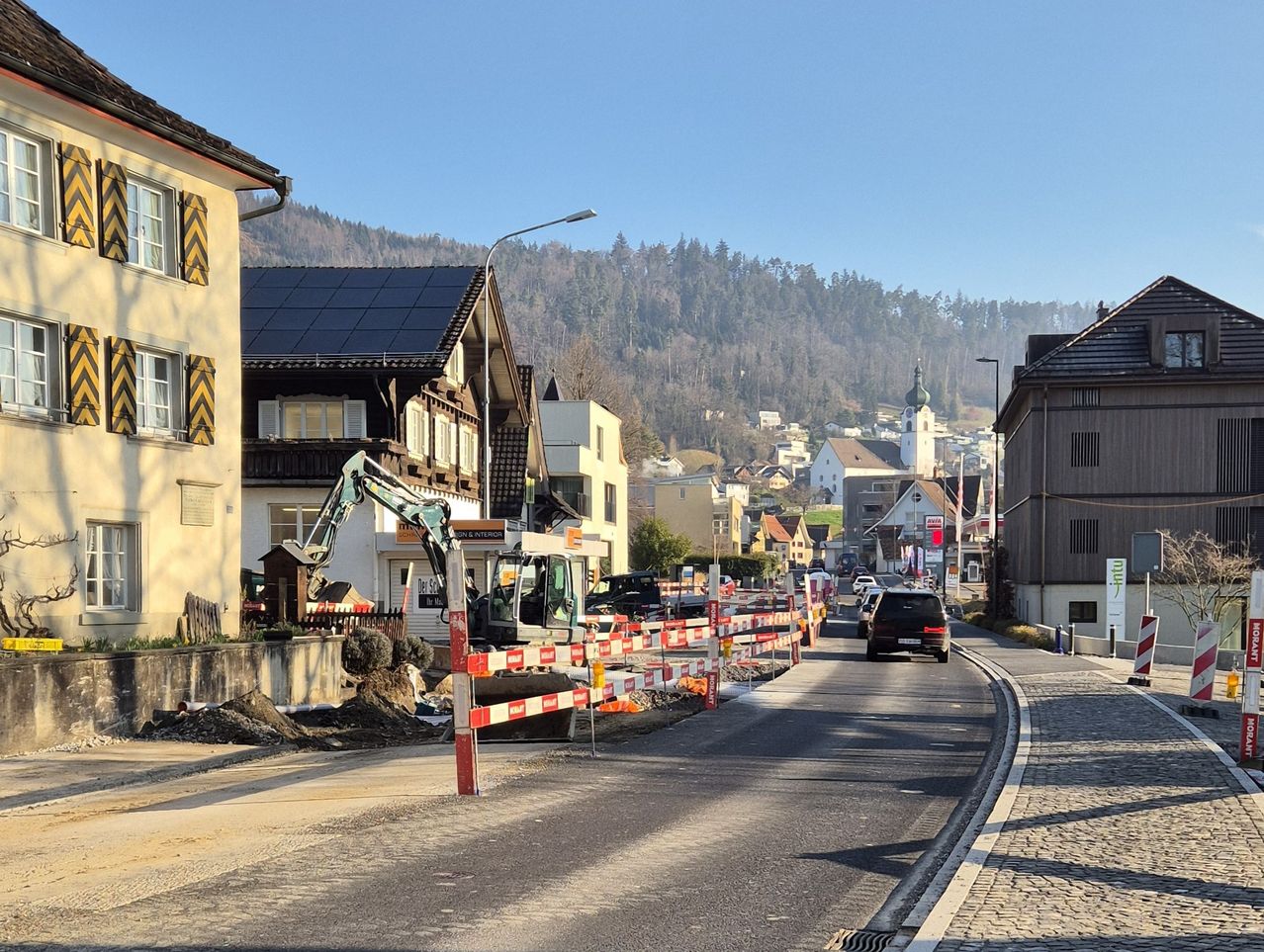 Eine Straße im Bau in einer Stadt mit einem vorbeifahrenden Auto, Gebäuden auf beiden Seiten und Bergen in der Ferne.