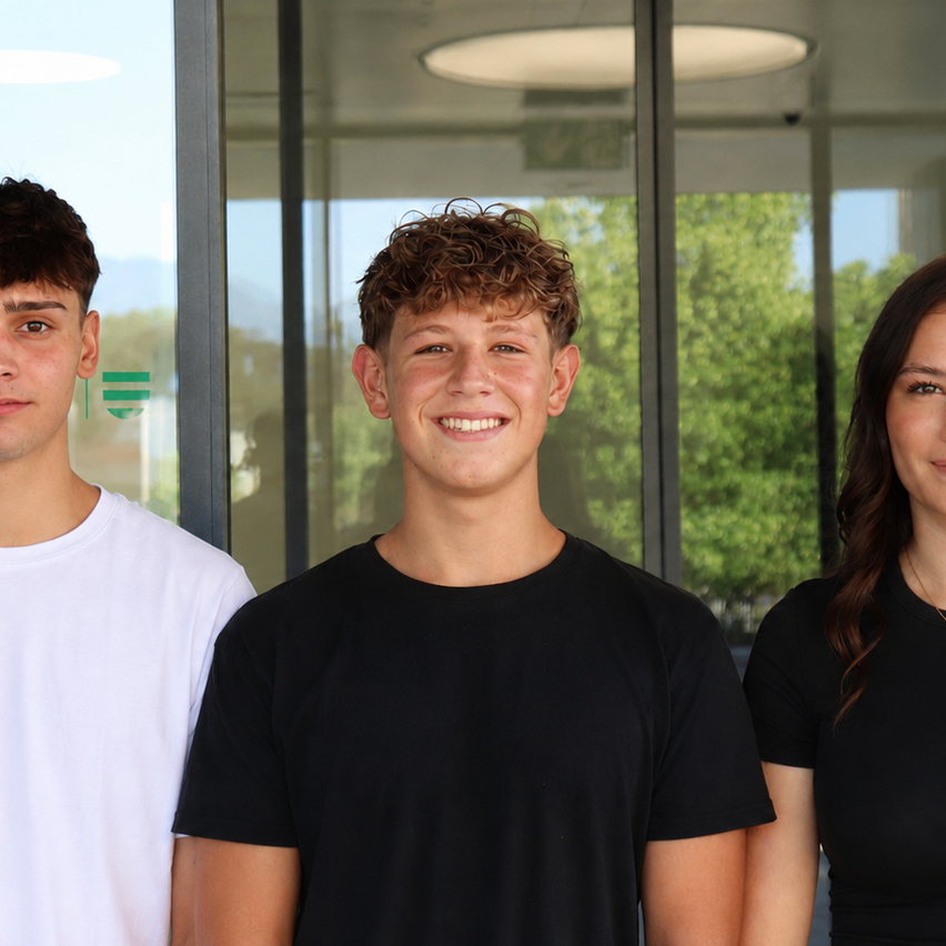 Three young individuals stand close together, smiling for the camera. The boy on the left wears a white shirt, the middle one a black shirt, and the girl on the right also wears a black shirt. They are in front of a glass wall with a logo and trees visible outside.