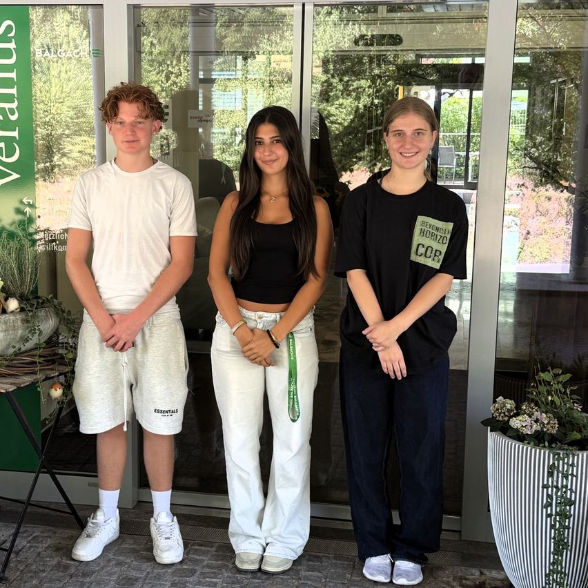 Three young people stand in front of a glass door with a sign that reads 'Veranius'. The boy on the left wears a white t-shirt and shorts, the girl in the middle wears a black tank top and white pants, and the girl on the right wears a black t-shirt with a green patch.