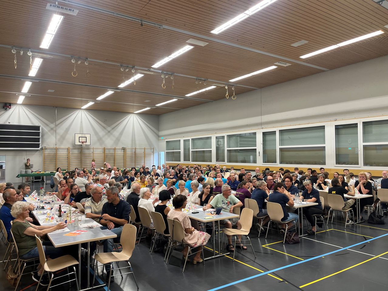 A large group of people are seated around tables in a hall. The hall has wooden ceilings, fluorescent lights, and glass windows. A basketball hoop is mounted on the wall.
