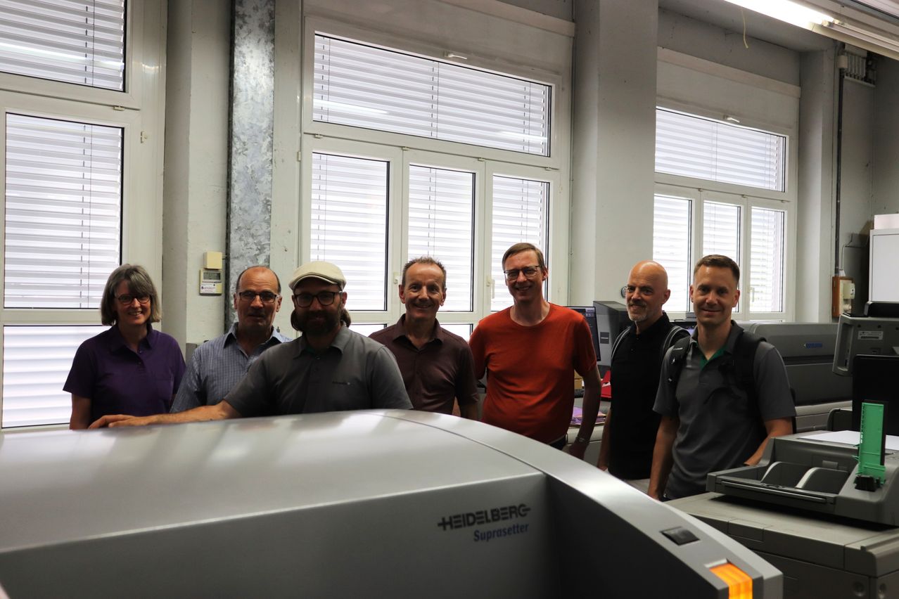 Eine Gruppe von Männern steht um eine große graue Heidelberg Suprasetter-Maschine und lächelt für das Foto.