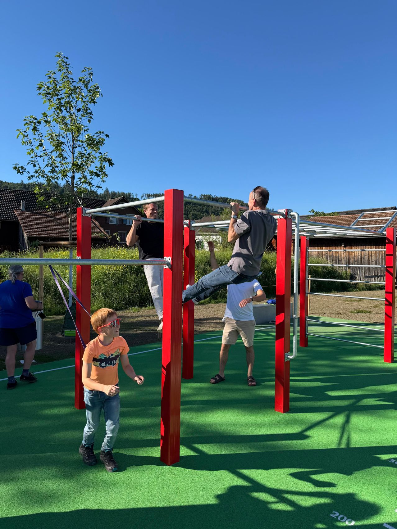 Drei Männer und ein Junge machen Klimmzüge an roten Stangen in einem Park. Zwei Männer hängen an den Stangen, während der Junge steht und zusieht.