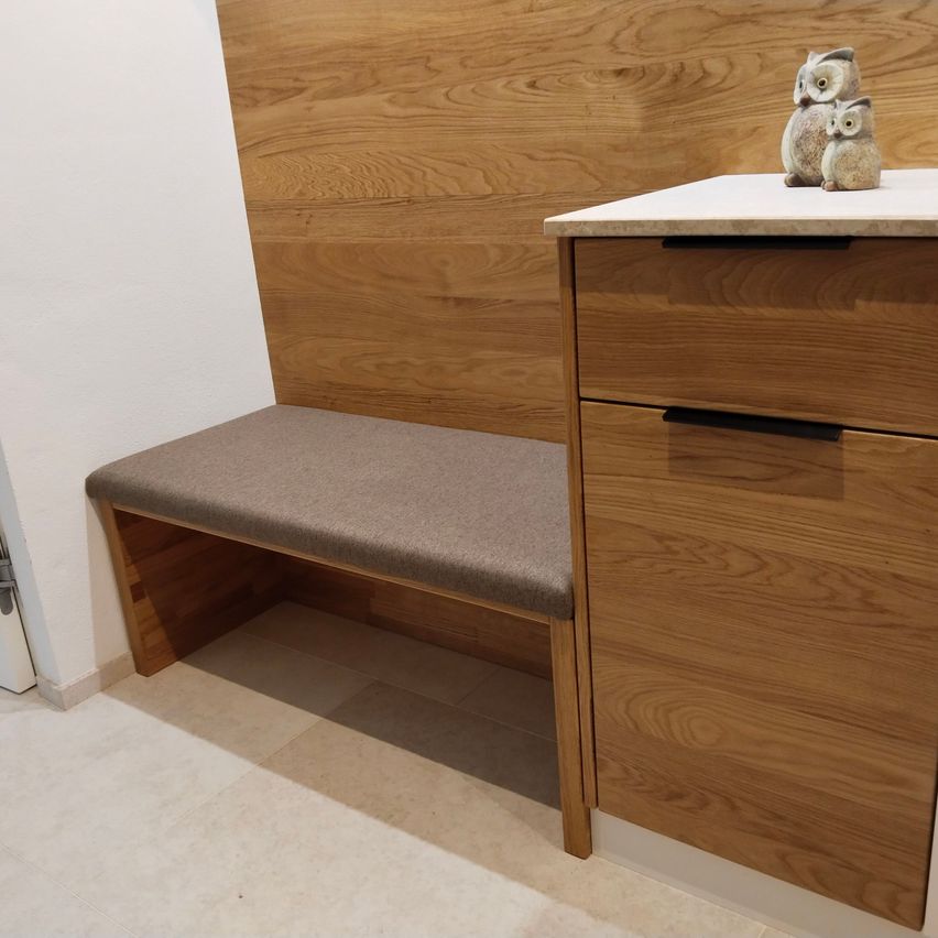 Eine Holzbank mit gepolsterter Sitzfläche steht an einer Holzwand. Ein weißer Arbeitstisch mit einer Holzschublade befindet sich neben der Bank, mit einer Figur von zwei Eulen darauf.