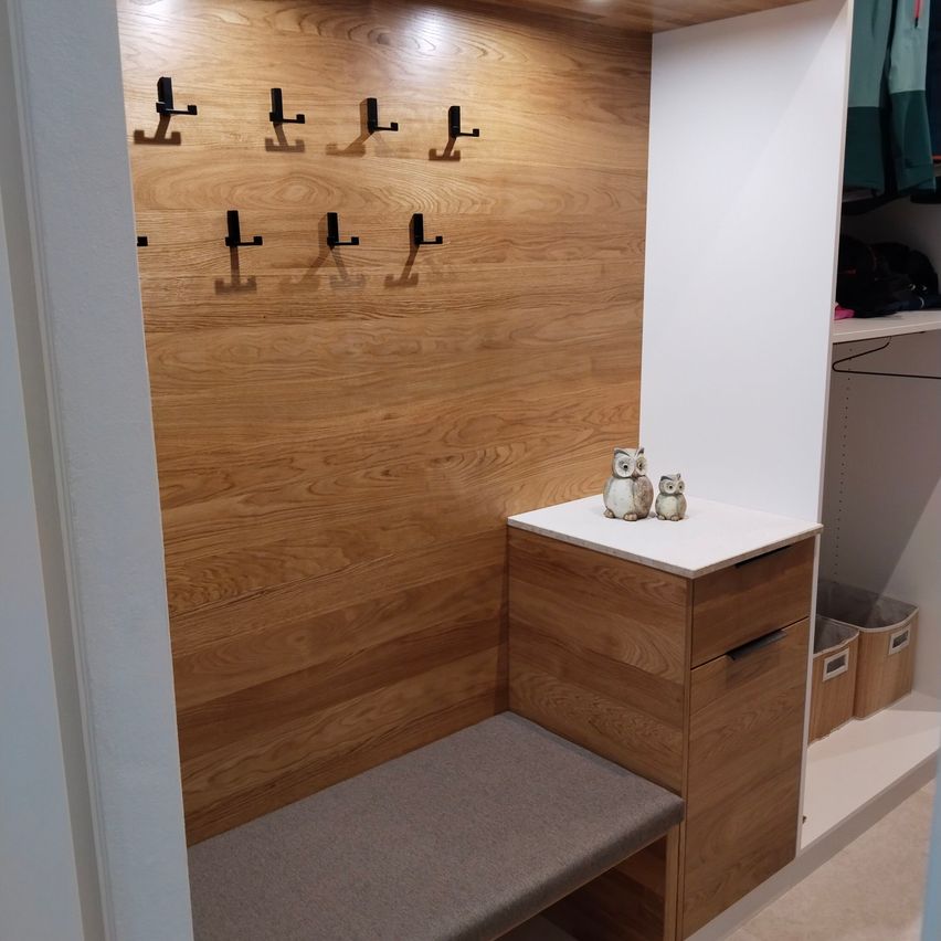 Ein modernes Interieur mit einer Holzwand, einer Bank und einem kleinen Holzschrank mit zwei Eulenfiguren darauf. Neben dem Schrank befindet sich ein Kleiderschrank mit Kleidern an Kleiderbügeln und Aufbewahrungskörben.