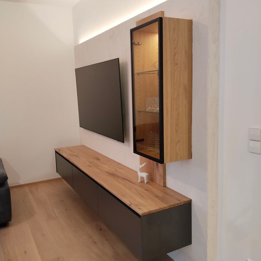 A modern living room features a flat-screen TV mounted above a wooden media unit with a glass display cabinet on the wall. The room has a wooden floor and white walls.