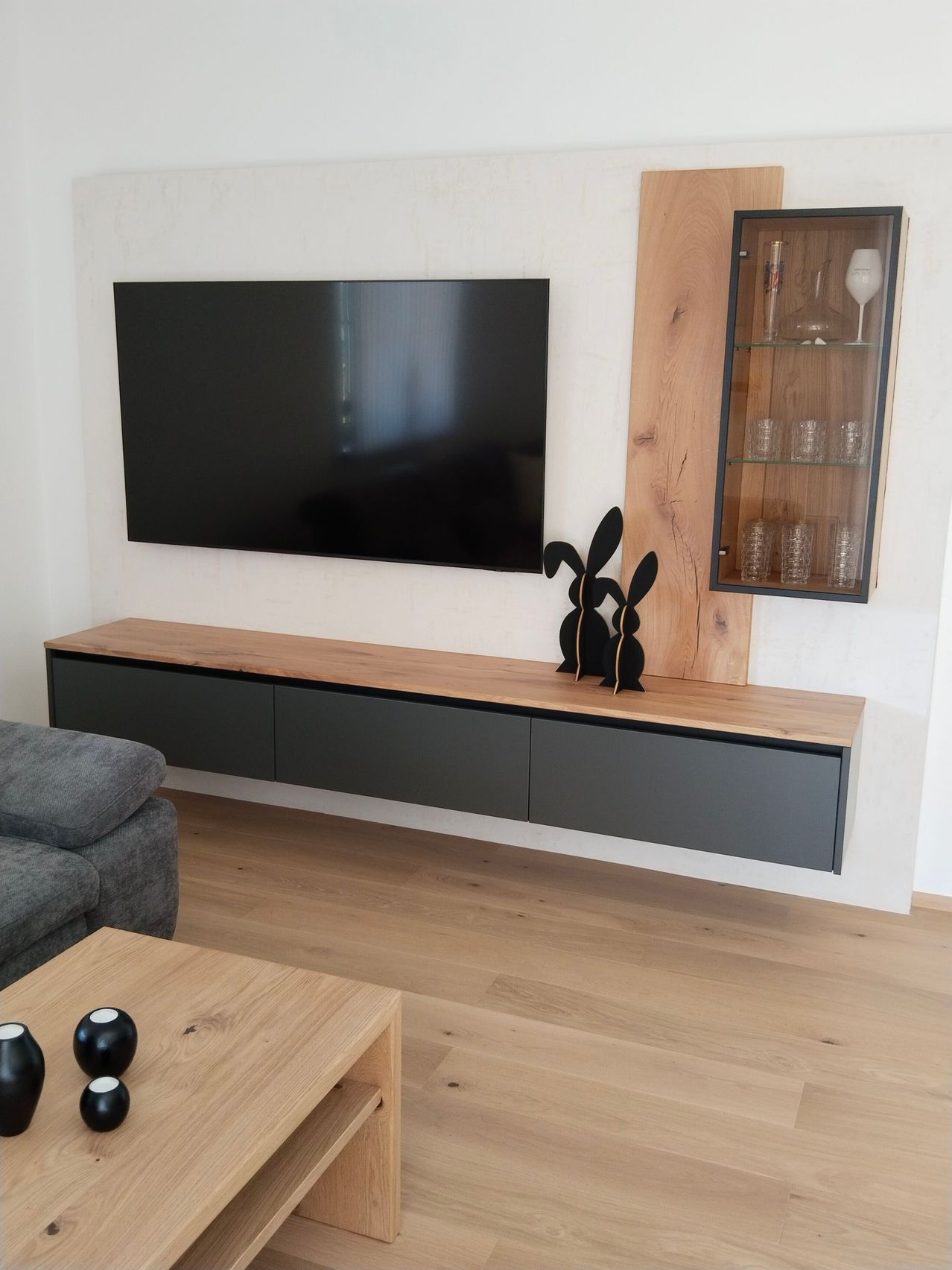 A modern living room with a large flat-screen TV mounted on the wall. The TV is turned off. Below it is a wooden entertainment center with a cabinet and a shelf with glass doors. There is a couch on the left.