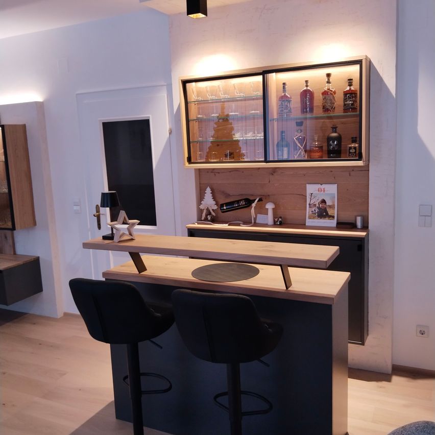 A modern kitchen bar with two chairs, a wooden counter, and a display cabinet with bottles and a magazine. A lamp and a Christmas decoration are on the counter.