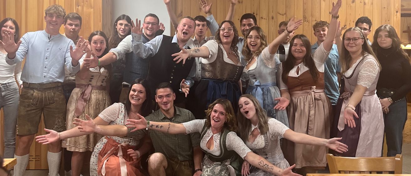 Eine Gruppe von Menschen in traditioneller Kleidung posiert für ein Foto in einem Raum mit Holzwänden. Einige stehen, andere knien und alle haben ihre Hände erhoben.
