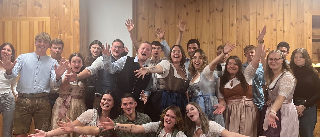 Eine Gruppe von Menschen in traditioneller Kleidung posiert für ein Foto in einem Raum mit Holzwänden. Einige stehen, andere knien und alle haben ihre Hände erhoben.